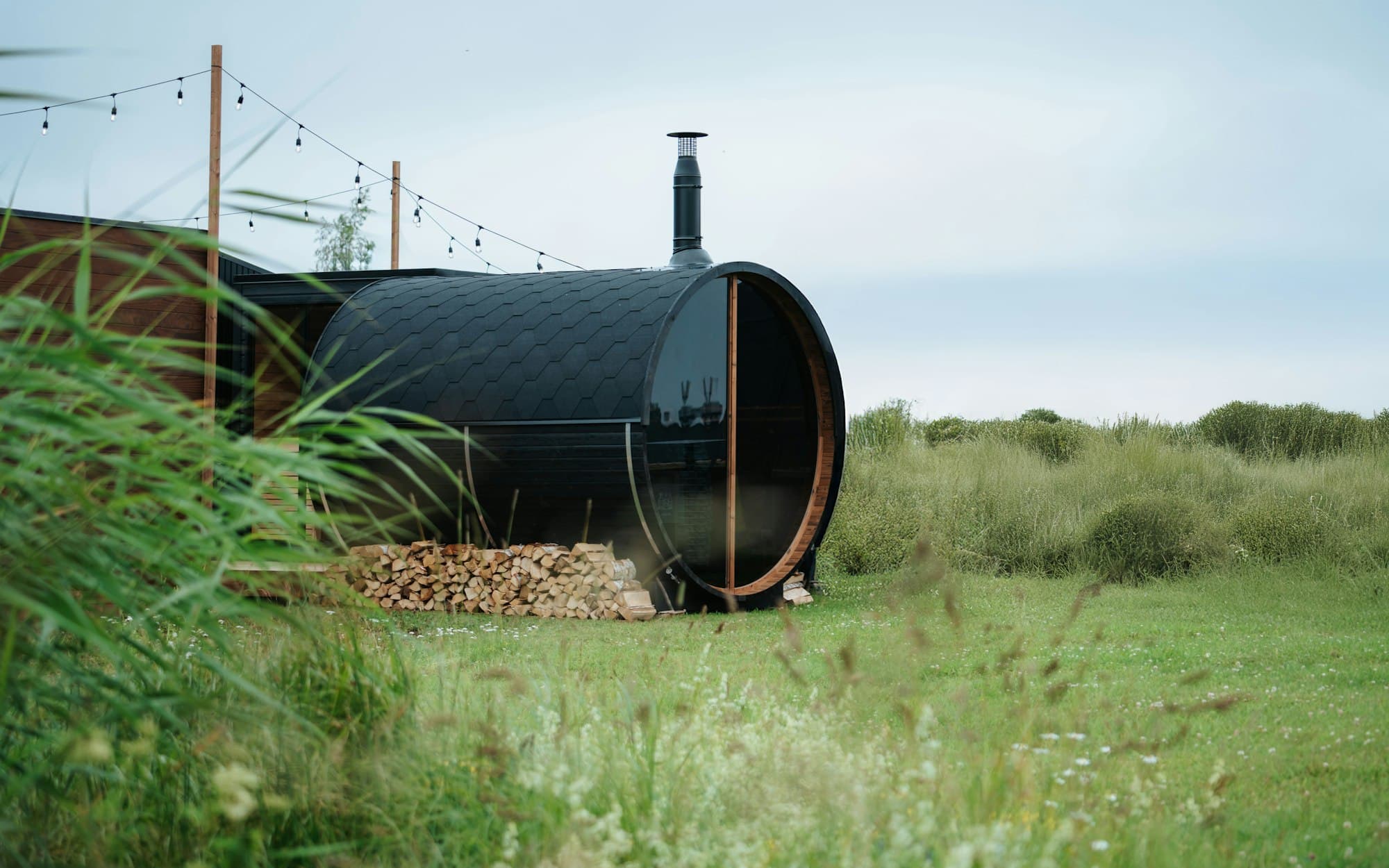 A barrel sauna with a chimney and a stack of firewood in a grassy field