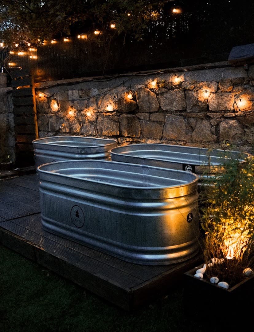 Three metal stock tanks sit on a dark wooden deck in front of a stone wall, illuminated by string lights.