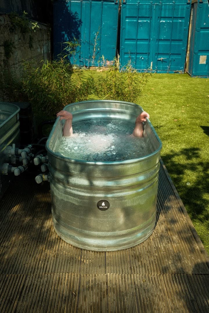 A person submerged in an oval metal ice bath on a wooden deck outdoors.