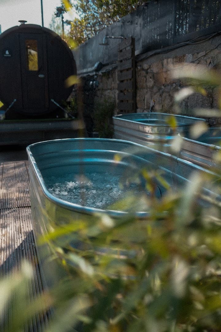 Two metal tubs filled with water sit outdoors next to a barrel sauna and a stone wall.