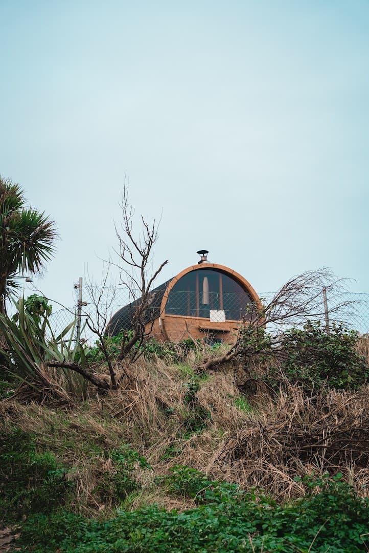 A barrel-shaped wooden sauna sits atop a grassy embankment beneath an overcast sky.