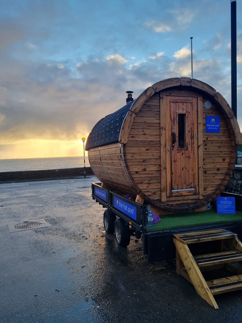 A mobile wooden barrel sauna is parked near the sea under a cloudy sunset sky.