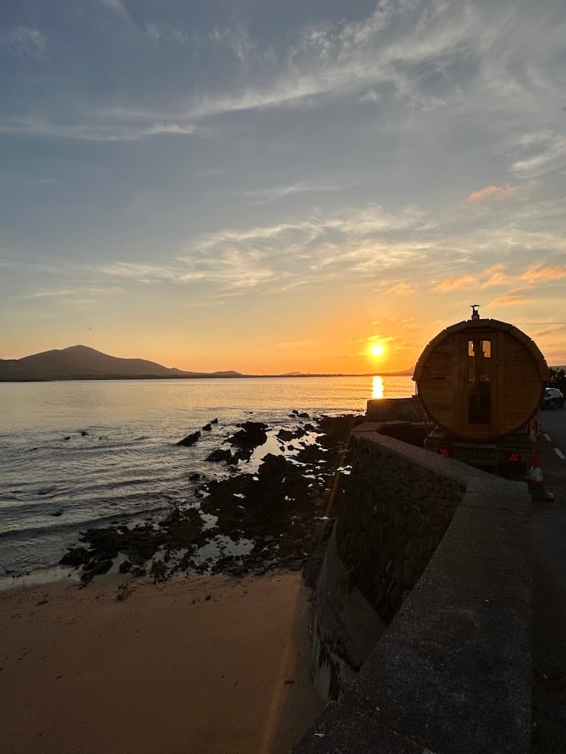 A barrel-shaped wooden sauna sits beside a coastal road overlooking a calm sea at sunset.