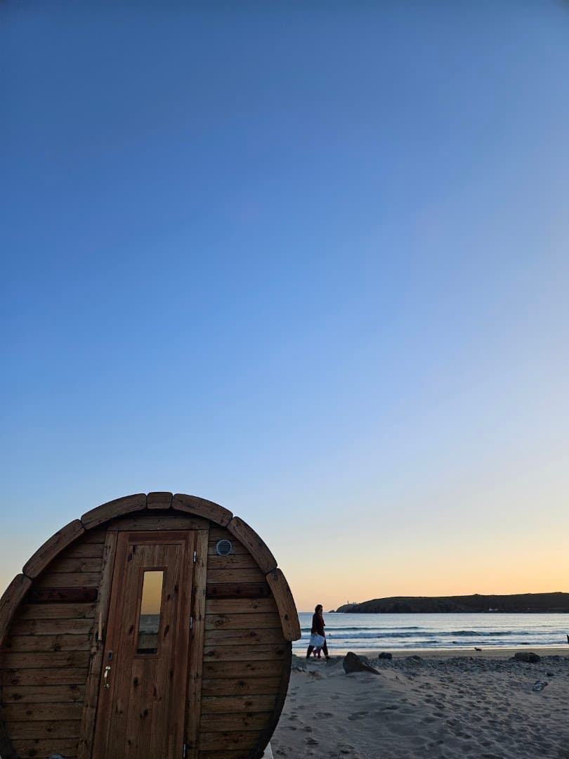A wooden barrel sauna on a sandy beach next to the sea at sunset.