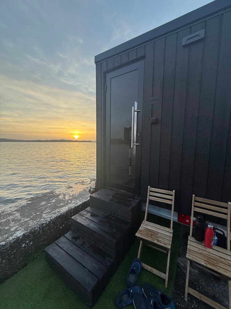 A dark wooden sauna stands on a concrete pier overlooking a lake at sunset.