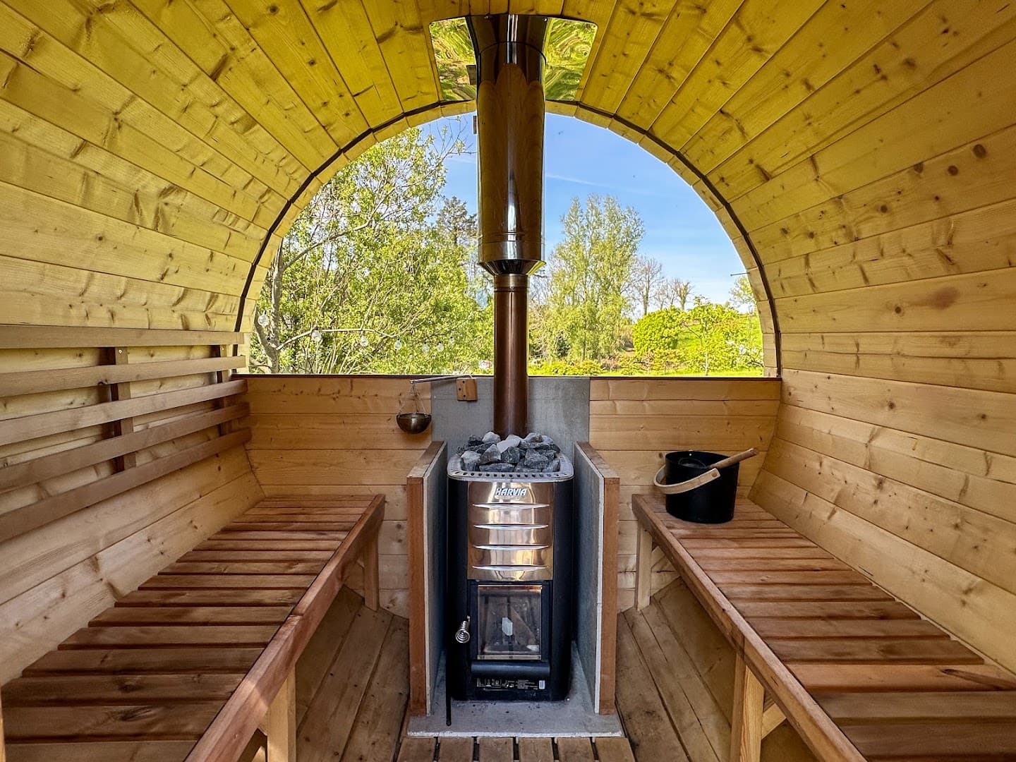 Interior view of a barrel sauna facing a wood-burning stove with a large arched window overlooking trees.