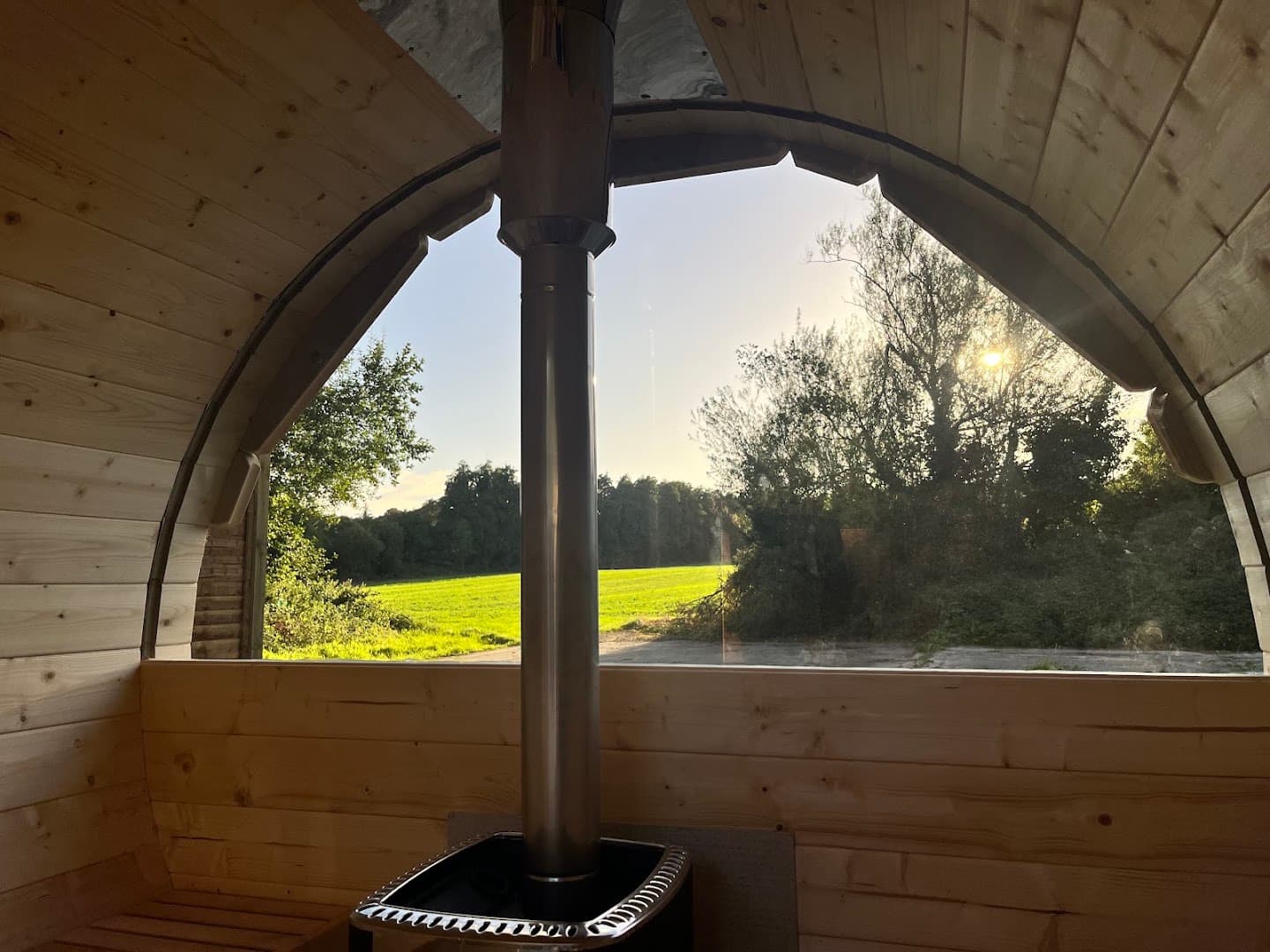 A view from inside a wooden sauna showing a metal stove, chimney, and a sunny green landscape outside.