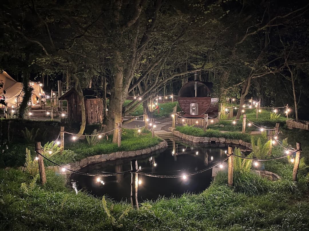 Illuminated barrel sauna in a lush, wooded clearing at night, with a small pond in the foreground.