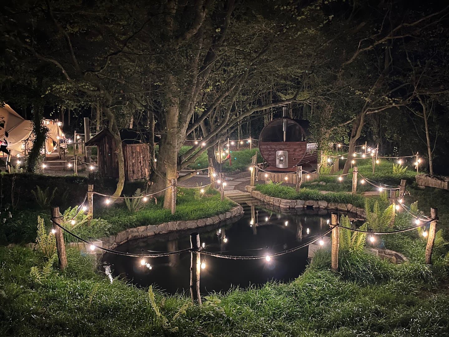 Illuminated barrel sauna in a lush, wooded clearing at night, with a small pond in the foreground.