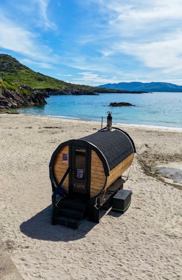 A barrel-shaped wooden sauna sits on a sandy beach next to the sea under a blue sky.