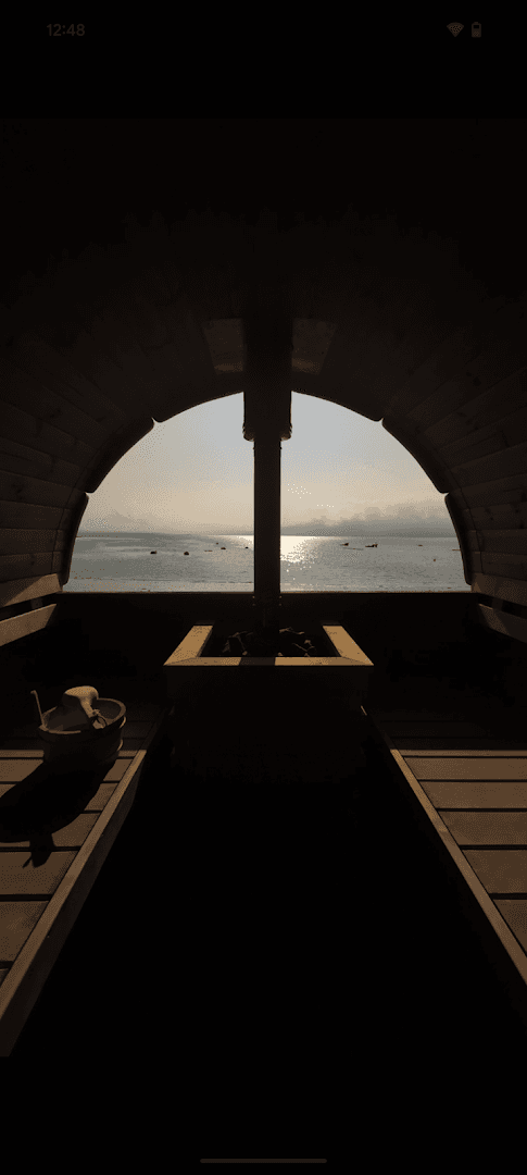 View of the sea through a large arched window from inside a wooden barrel sauna with a central stove.