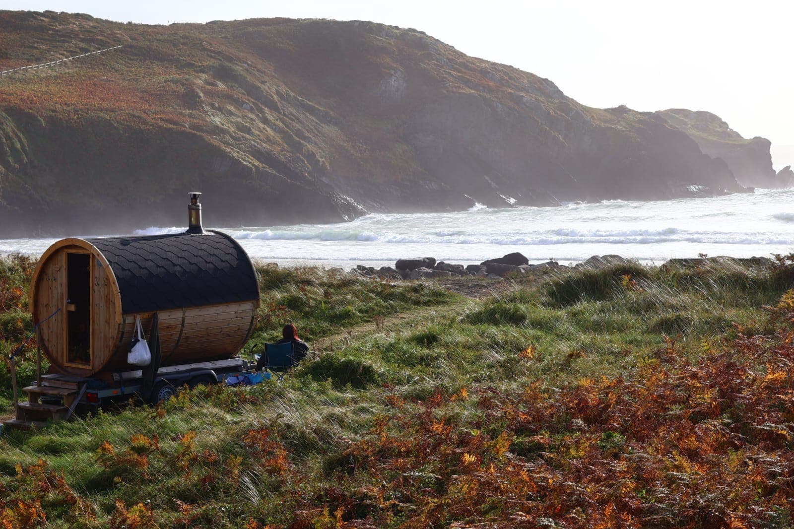A wooden barrel sauna on a trailer is parked on a grassy coast overlooking the ocean and rocky hills.