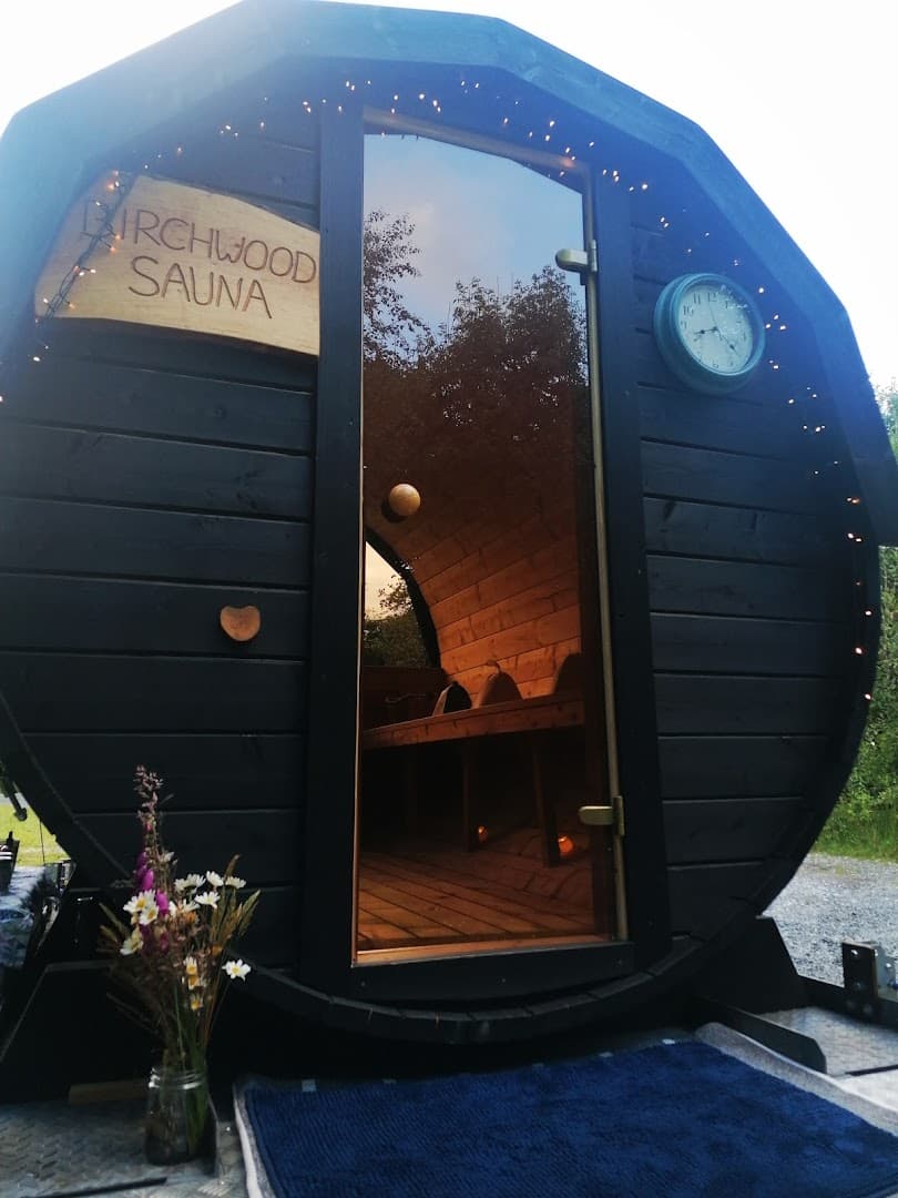 Black wooden barrel sauna with a glass door, a sign reading Birchwood Sauna, and a clock on the wall.