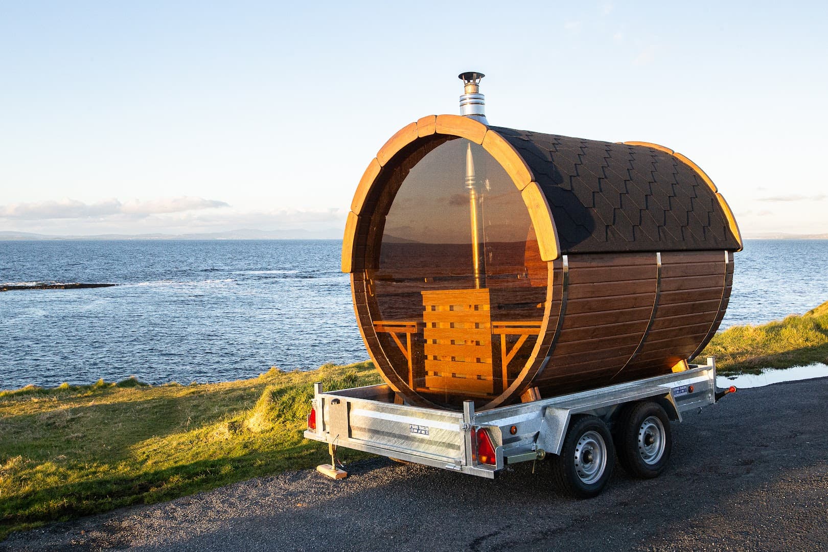 A wooden barrel sauna on a trailer sits by the sea with a view of the water and coastline.
