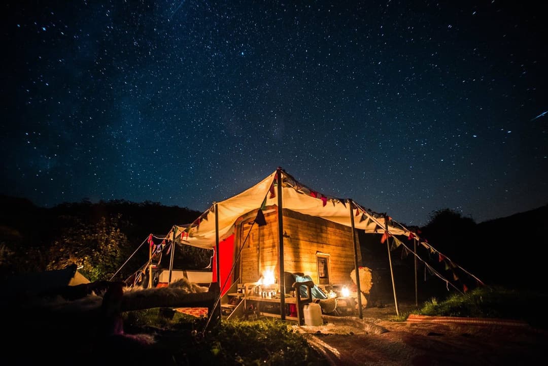 A wooden sauna lit from within stands outdoors under a vast star-filled night sky.