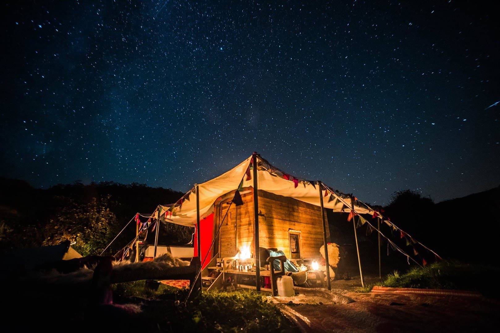 A wooden sauna lit from within stands outdoors under a vast star-filled night sky.