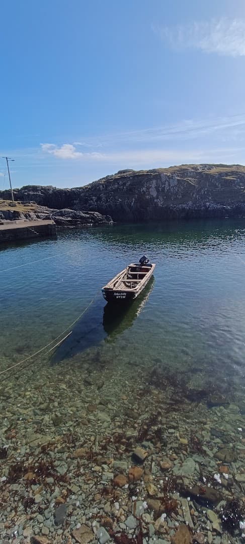 A small boat is moored in clear shallow water near a rocky coast under a bright blue sky.