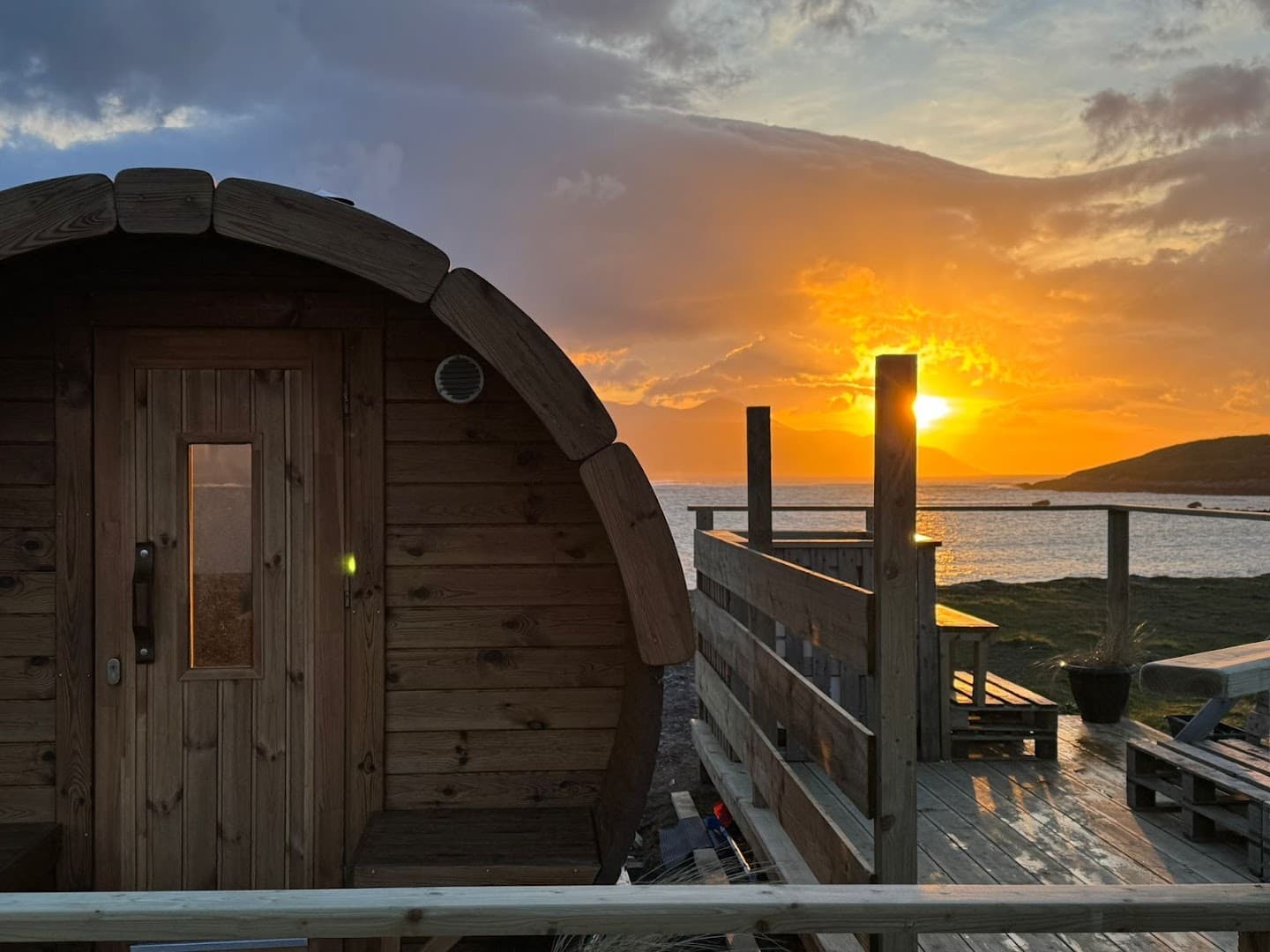 A wooden barrel sauna stands on a deck by the sea during a golden sunset.
