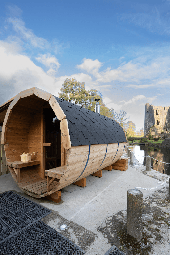 An outdoor wooden barrel sauna stands on a concrete platform near a body of water and a castle.