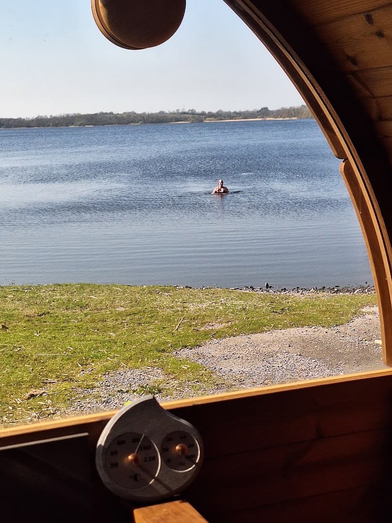 Looking through a wooden sauna window, a person is seen swimming in the calm lake outside.