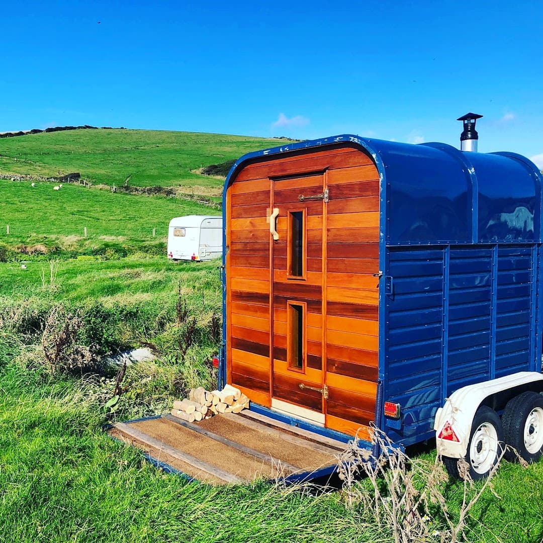 A wooden-paneled sauna converted from a blue trailer sits in a grassy field under a blue sky, with a small caravan in the distance.