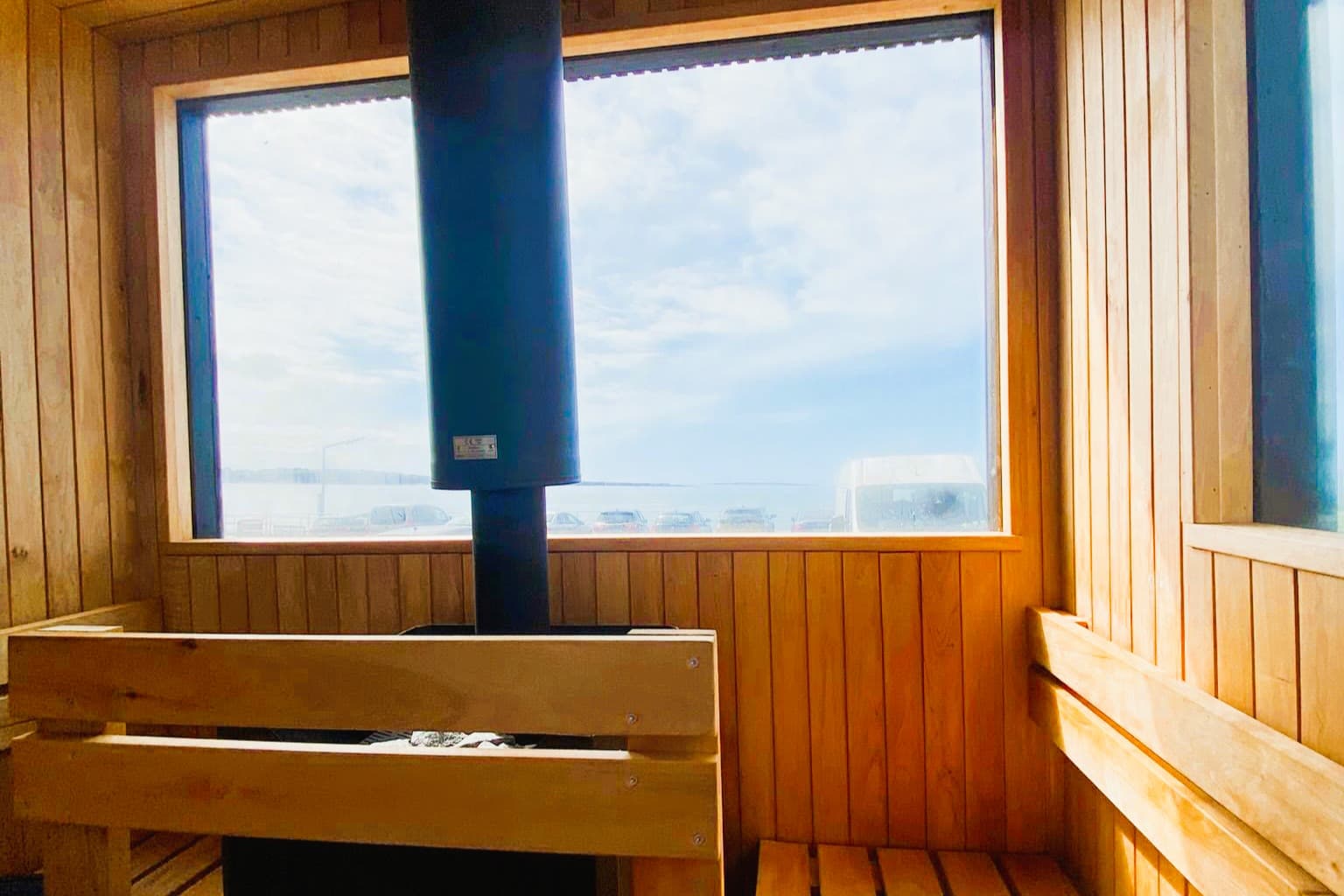 Inside a wooden sauna with a large window overlooking the sea, a stove and wooden benches are visible.