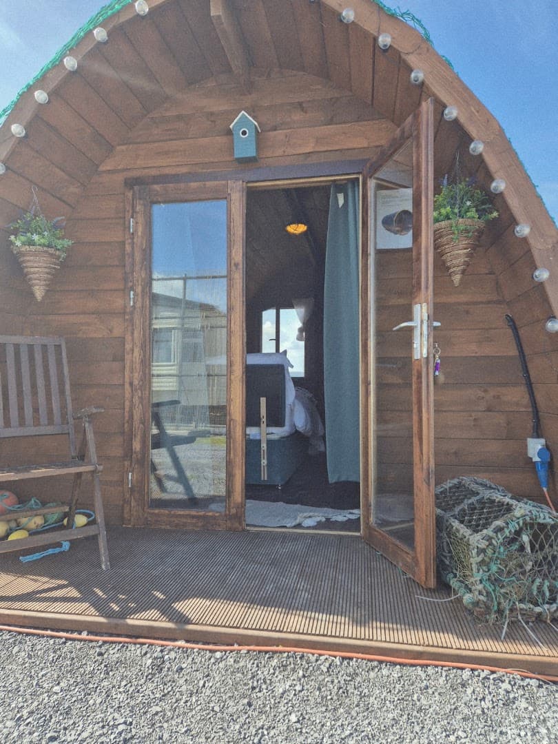 The wooden barrel sauna structure features a small porch with an open glass door, showing a bed inside.
