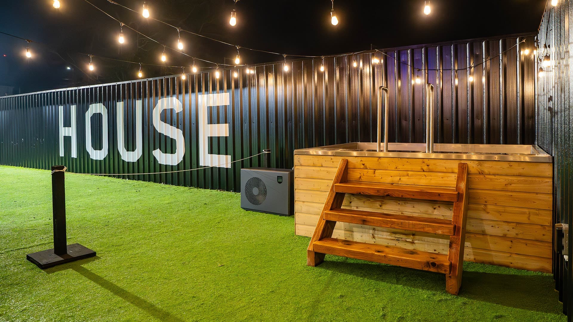 Outdoor sauna structure with wooden steps and railings against a corrugated wall on artificial grass at night.