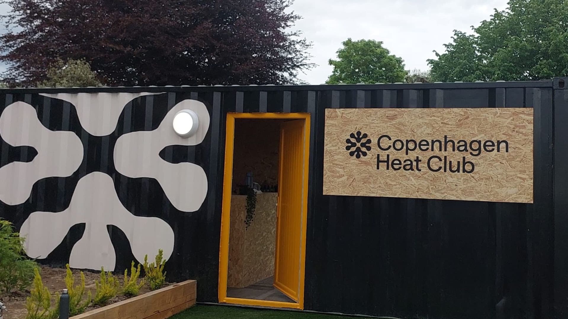 Black shipping container converted into the Copenhagen Heat Club with a yellow doorway, round light, and sign.