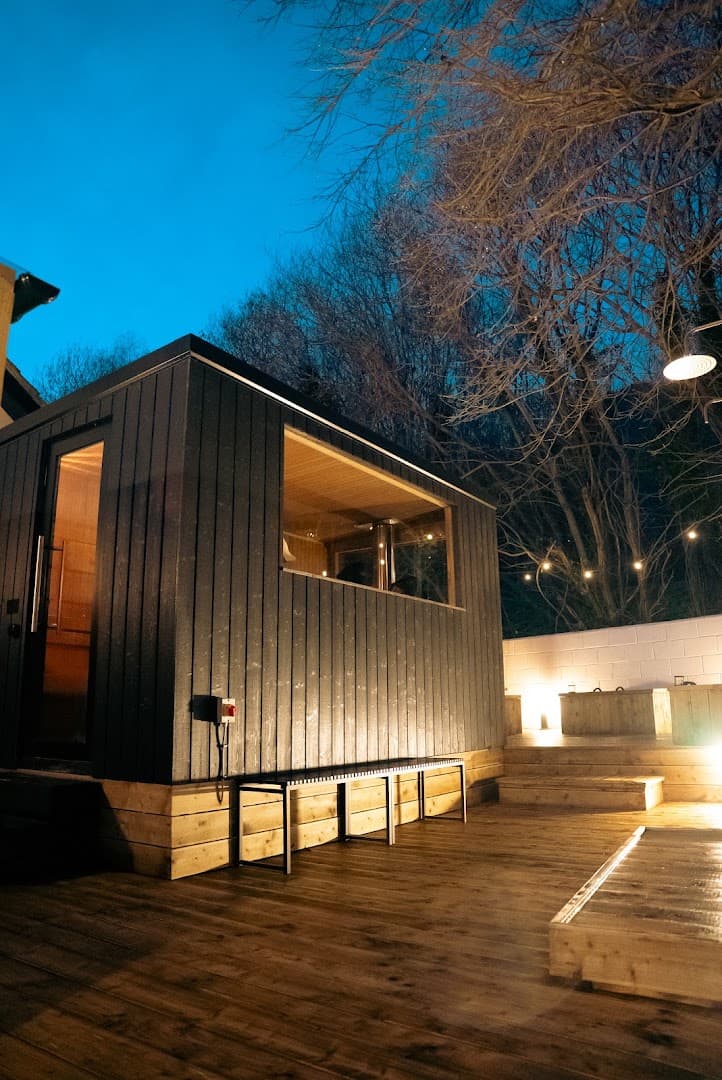 Exterior of a black wooden sauna with a large window and lit interior on a wooden deck.
