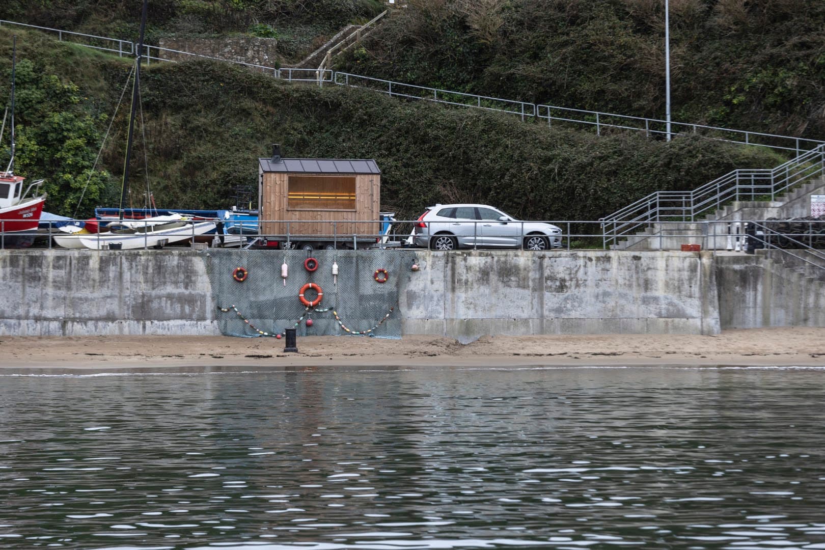 A wooden sauna cabin stands on a concrete pier next to a silver car and the sea.