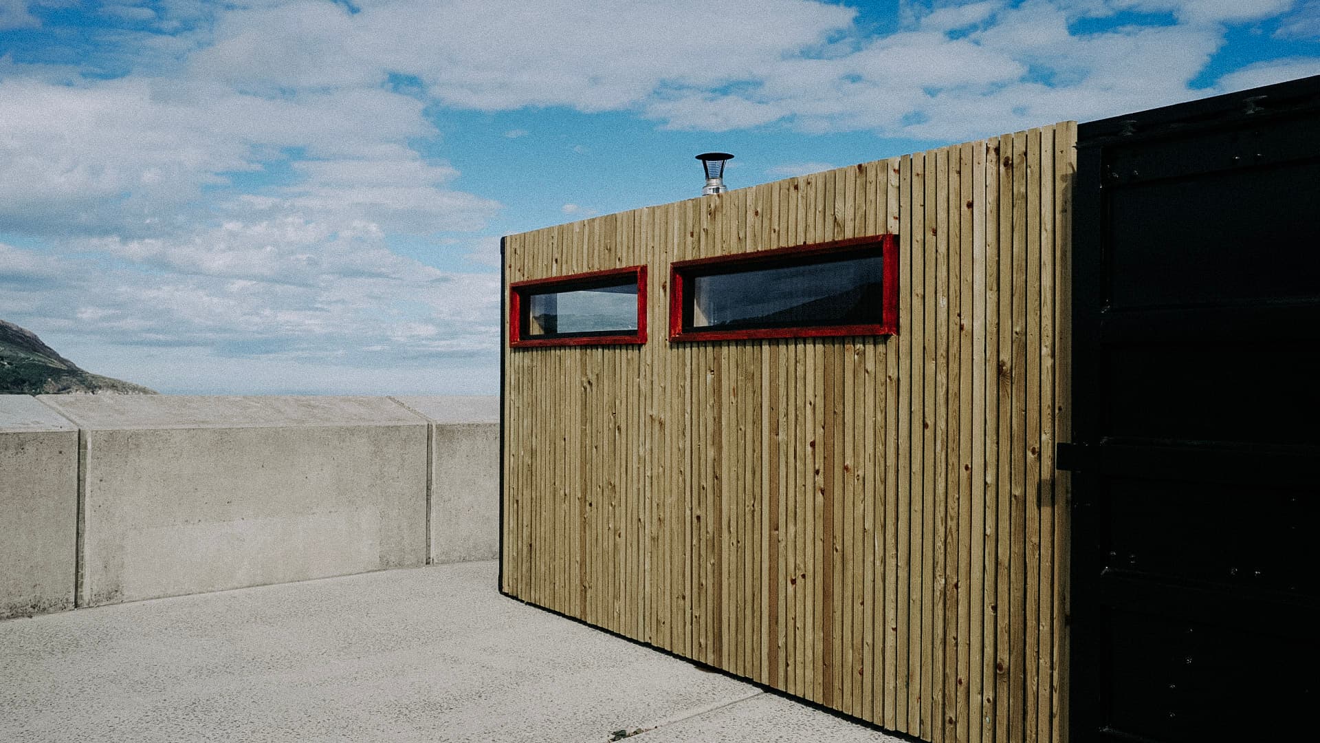 A wood-clad sauna structure with two red-framed rectangular windows stands on a concrete pier against a cloudy blue sky.