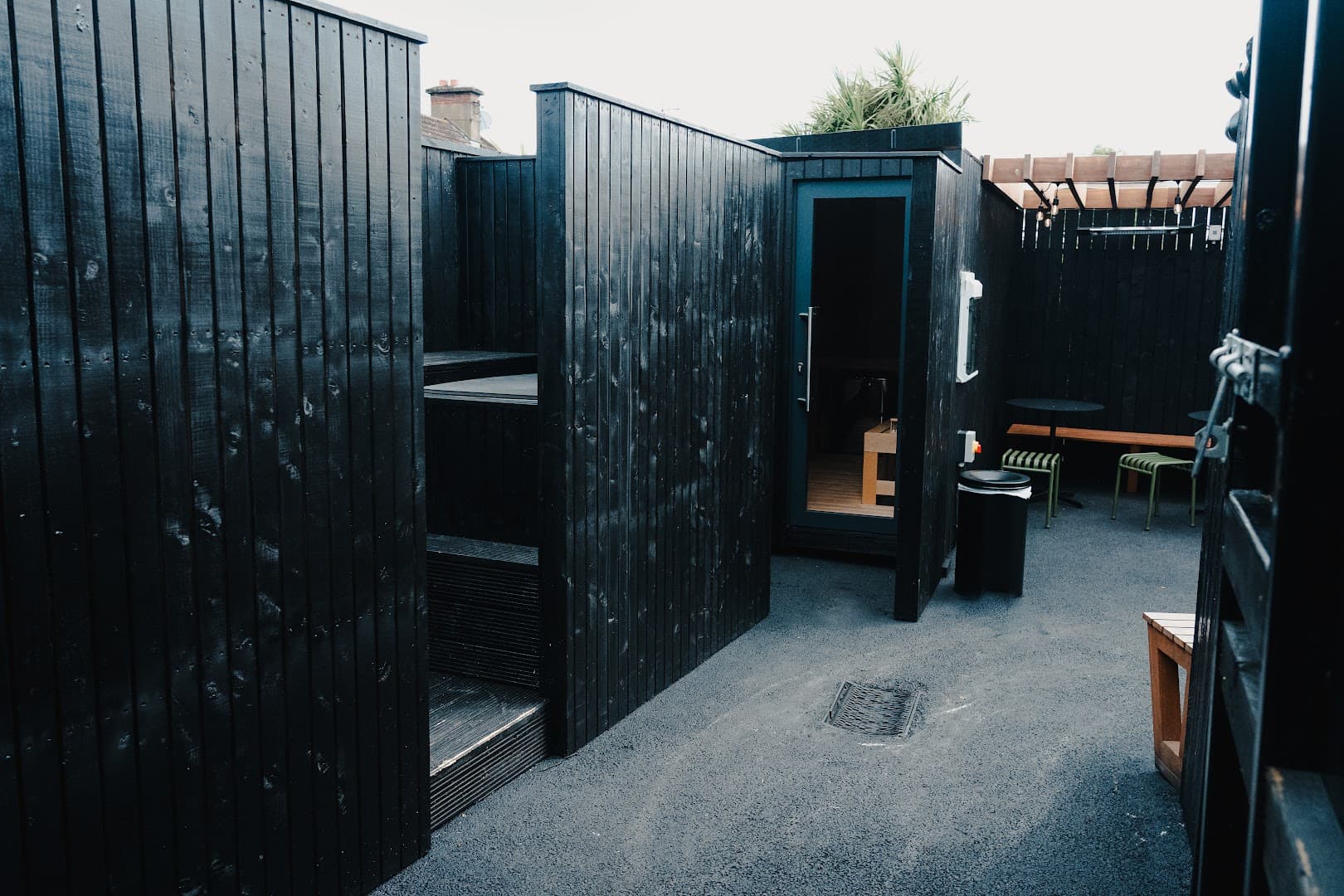 Black wooden sauna structures stand outdoors on a paved ground with seating in the background.
