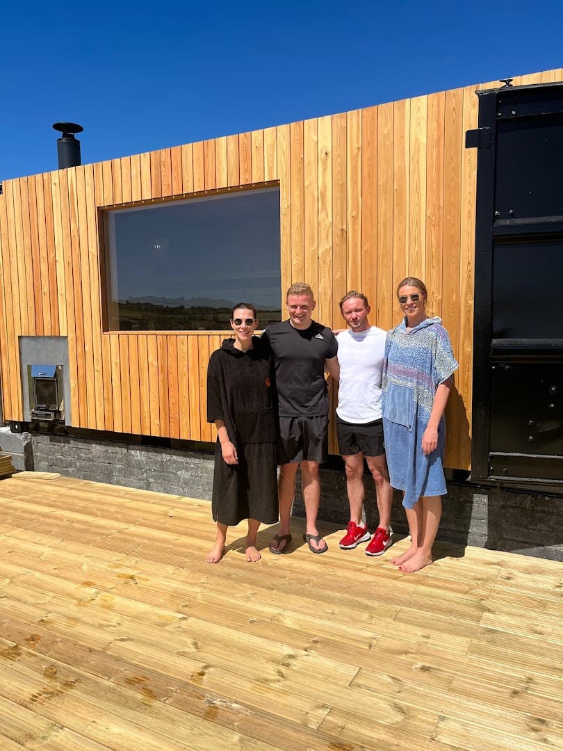 Four people stand smiling on a wooden deck in front of a wooden sauna structure under a bright blue sky.