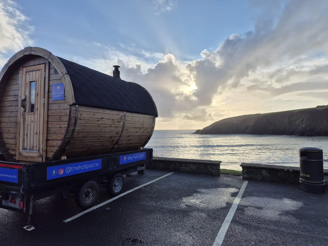 A wooden barrel sauna on a trailer sits by Kilmurrin Cove overlooking the sea under a cloudy sky.