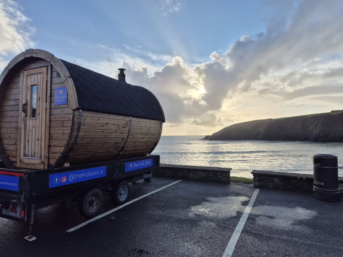 A wooden barrel sauna on a trailer sits by Kilmurrin Cove overlooking the sea under a cloudy sky.