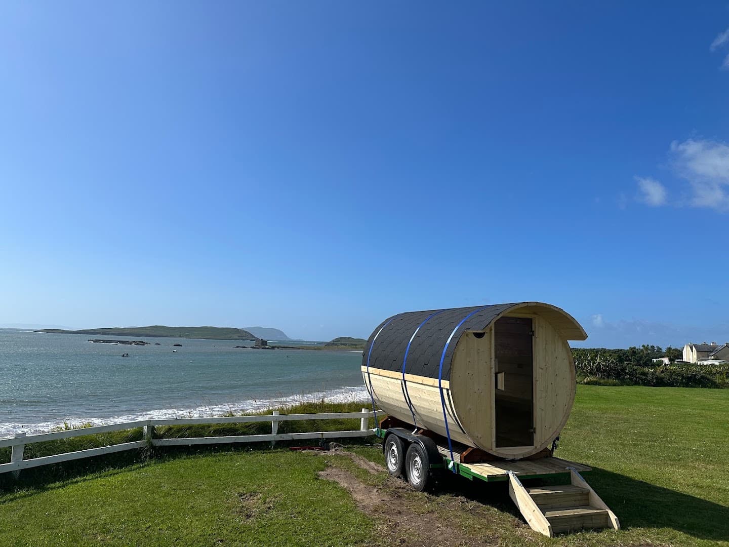 A cylindrical wooden sauna on a trailer sits on a grassy cliff overlooking the sea under a clear blue sky.
