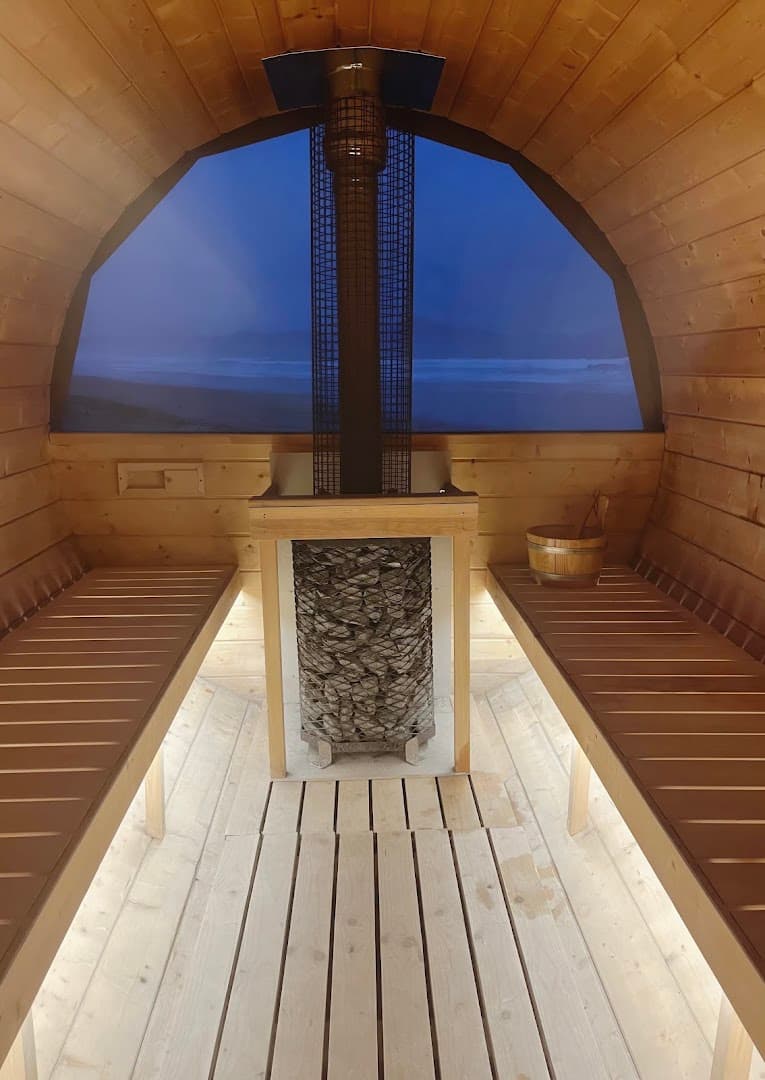 Interior of a wooden barrel sauna overlooking the sea through a large window, with a central stone heater.