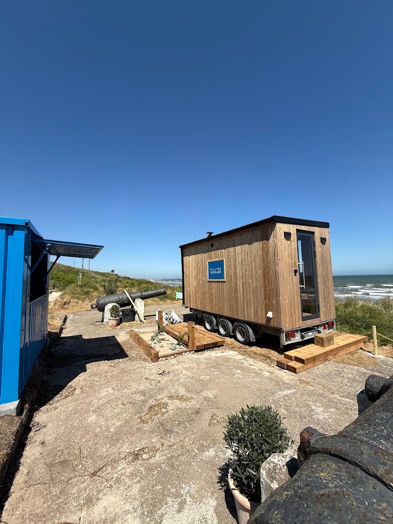 A wooden sauna on wheels is parked on a concrete area near the sea under a clear sky.