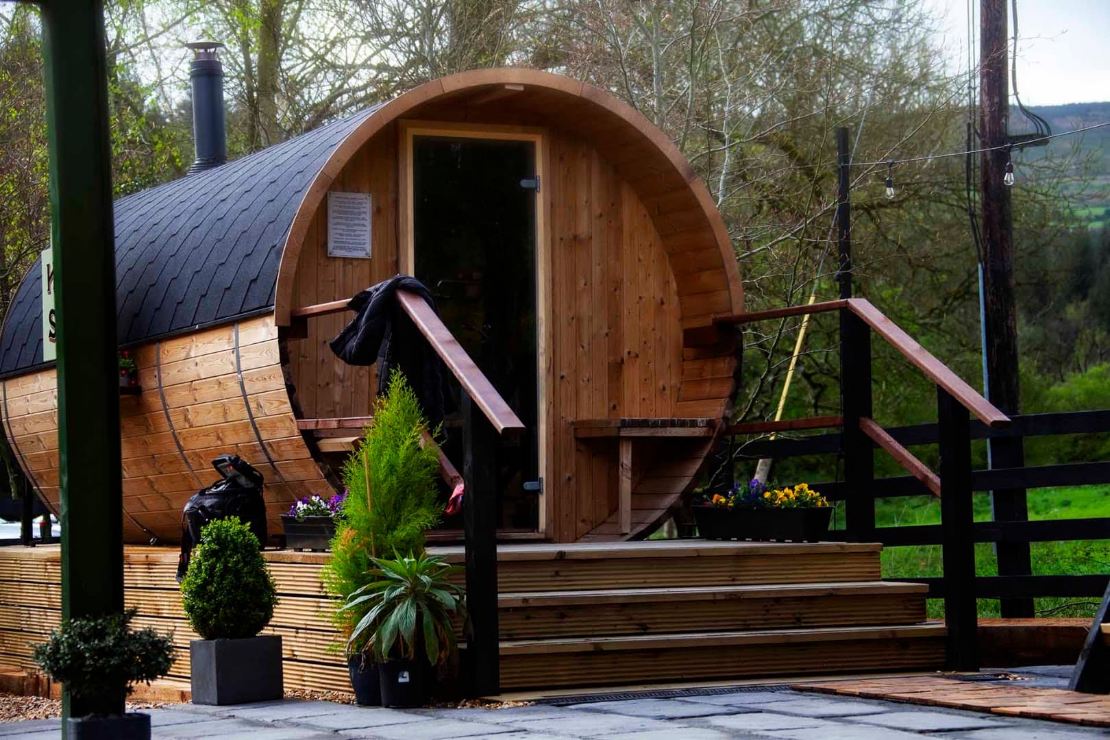 A wooden barrel-shaped sauna stands on a deck with stairs, surrounded by greenery and trees.