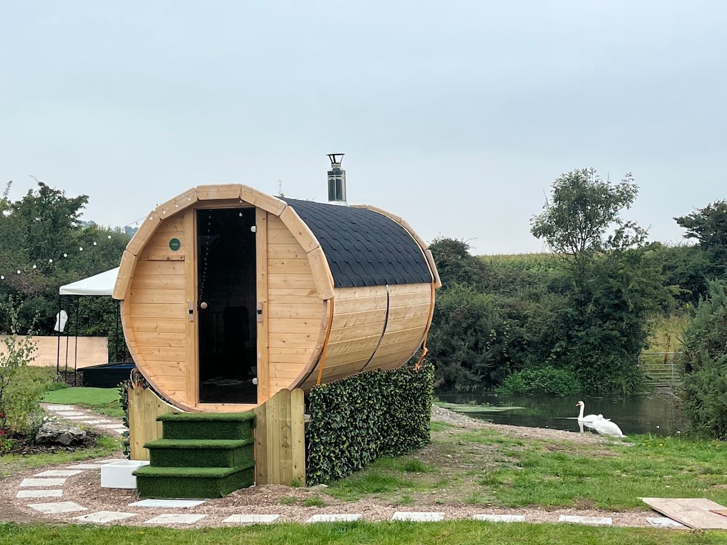 A wooden barrel sauna with a black shingle roof stands outdoors near a pond.