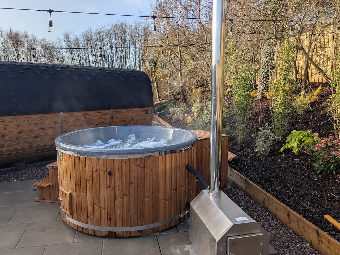A wooden hot tub with a metal stove stands on a paved patio beside a barrel sauna outdoors.