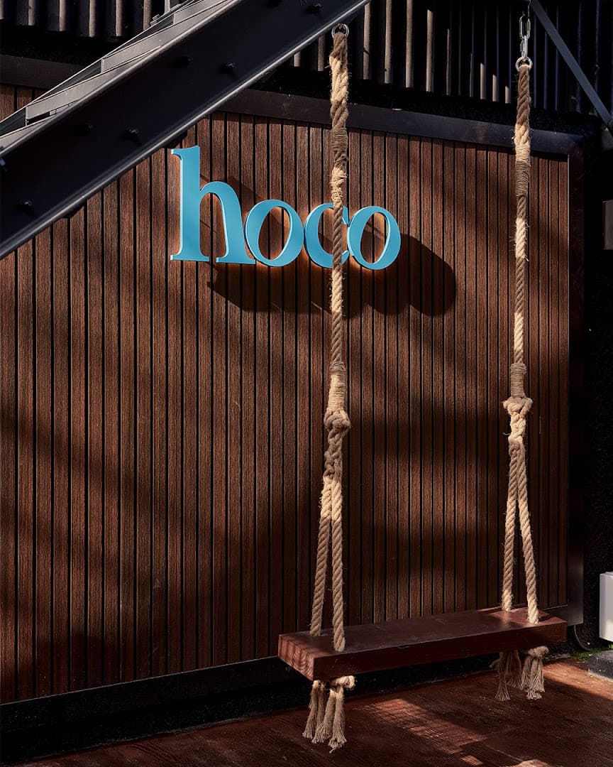 A wooden swing hangs in front of a slatted brown wall with a turquoise "hoco" sign.