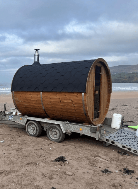 A wooden barrel sauna on a dual-axle trailer is parked on a sandy beach next to the sea under a cloudy sky.