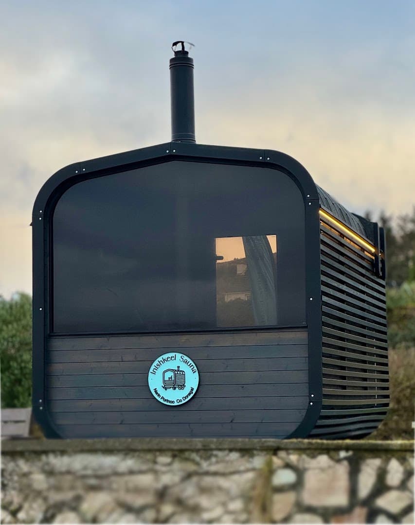 A dark wooden sauna building with a chimney stands outdoors against a cloudy sky.
