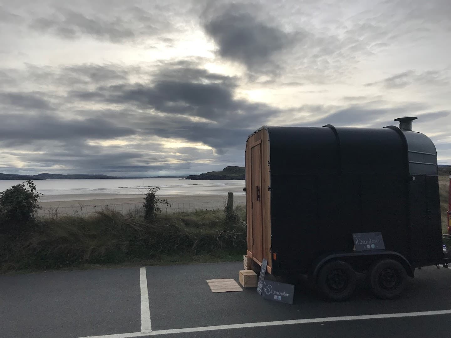 A black sauna trailer stands parked on asphalt overlooking a sandy beach and water under a cloudy sky.