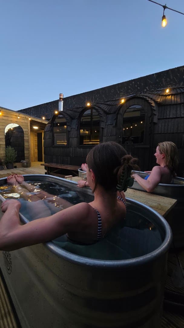 People relaxing in outdoor metal tubs in front of a dark wooden sauna structure under a twilight sky.