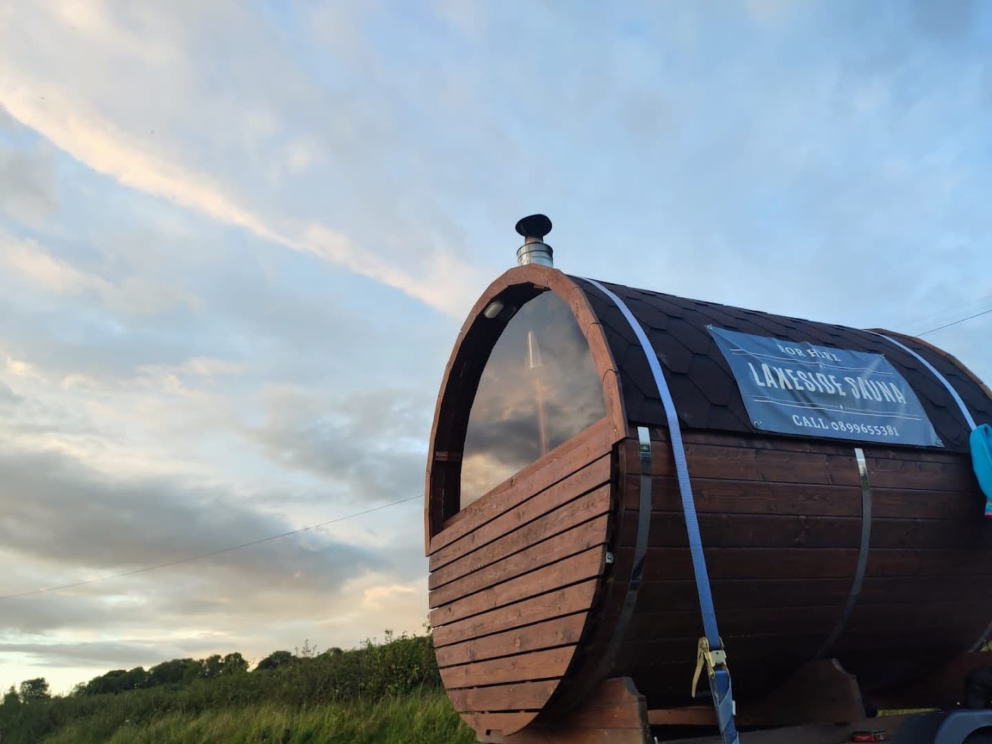 Wooden barrel sauna with a glass front and a metal chimney outdoors.