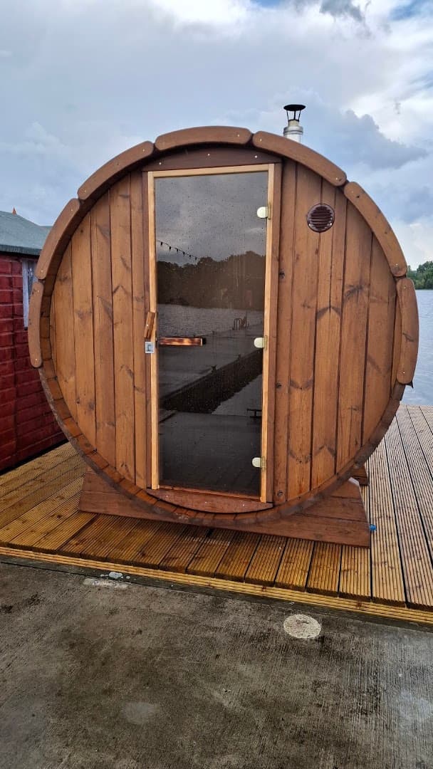 A barrel-shaped wooden sauna with a glass door stands on a wooden deck by a lake under a cloudy sky.