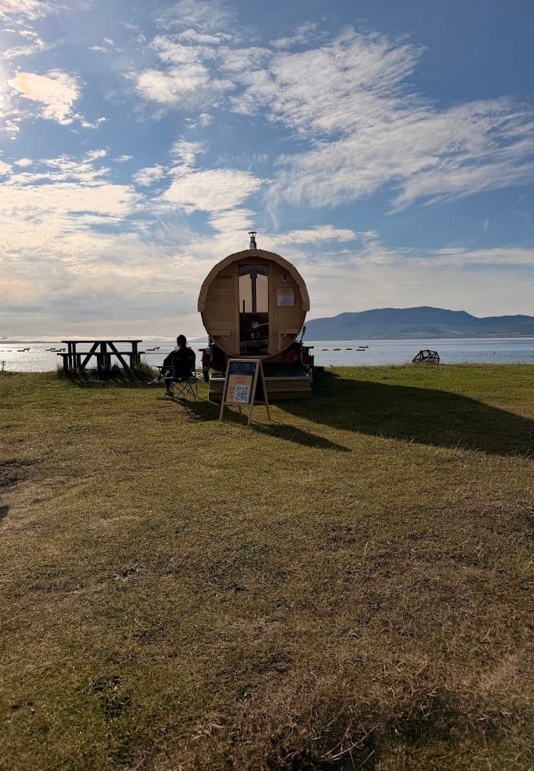 A barrel-shaped sauna on a grassy coastline overlooks the sea, with a person sitting nearby in a chair.